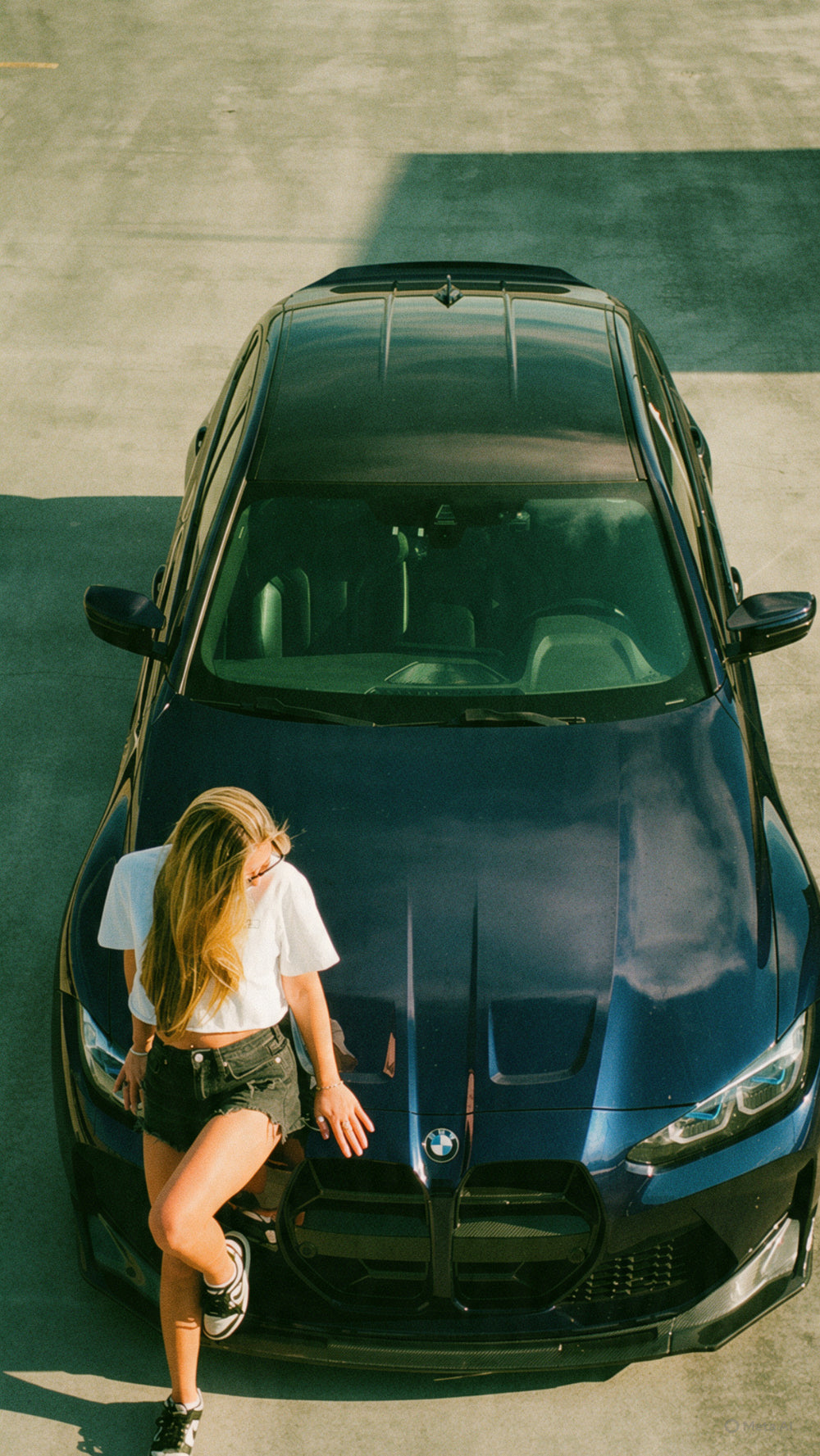 A woman in a white t-shirt and denim shorts leans against the front hood of a dark blue BMW sedan in an outdoor paved area.